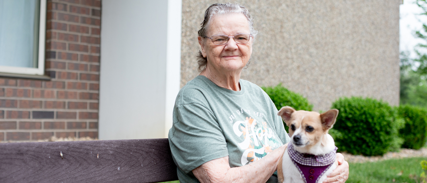 An elderly woman sitting outdoors in a light blue t-shirt with a dog in her arms