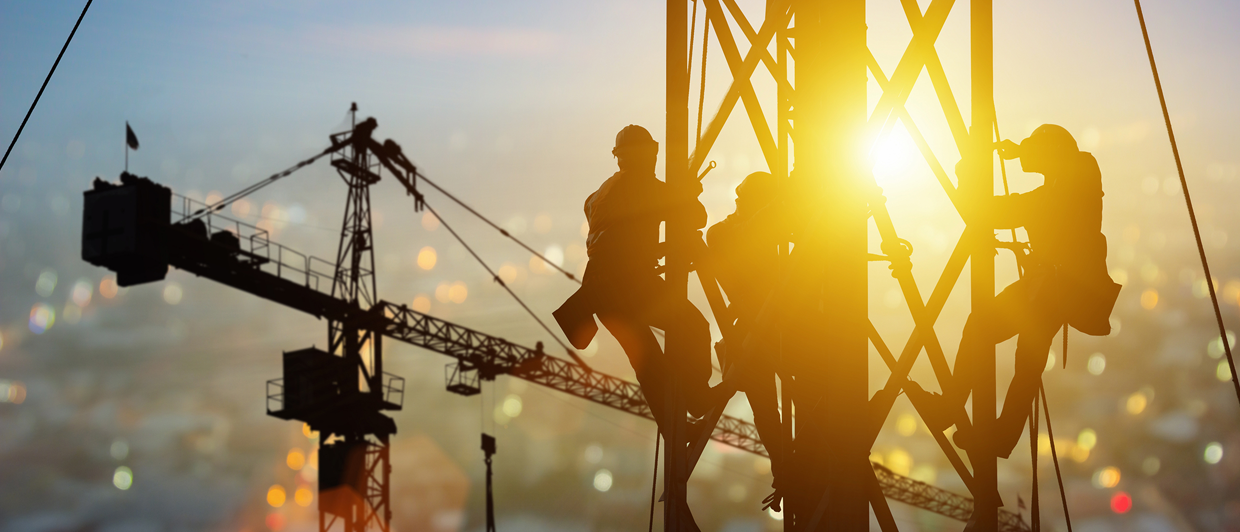 Silhouetted construction workers climb a tall metal structure at sunset, with cranes and blurred city lights in the background.